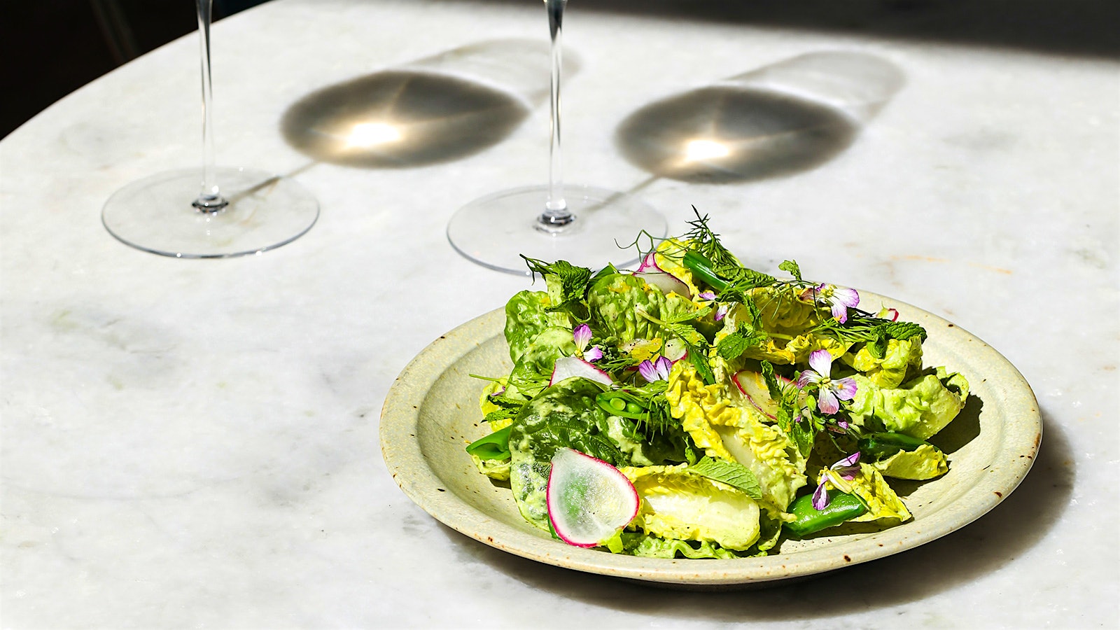 Spring salad with radishes and flowers on a table next to the shadow of two wine glasses.